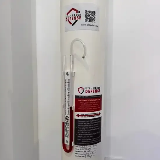Utah Radon Defense system label and QR code on a finished radon vent pipe, next to a manometer homeowners can read at a glance to confirm the system is working