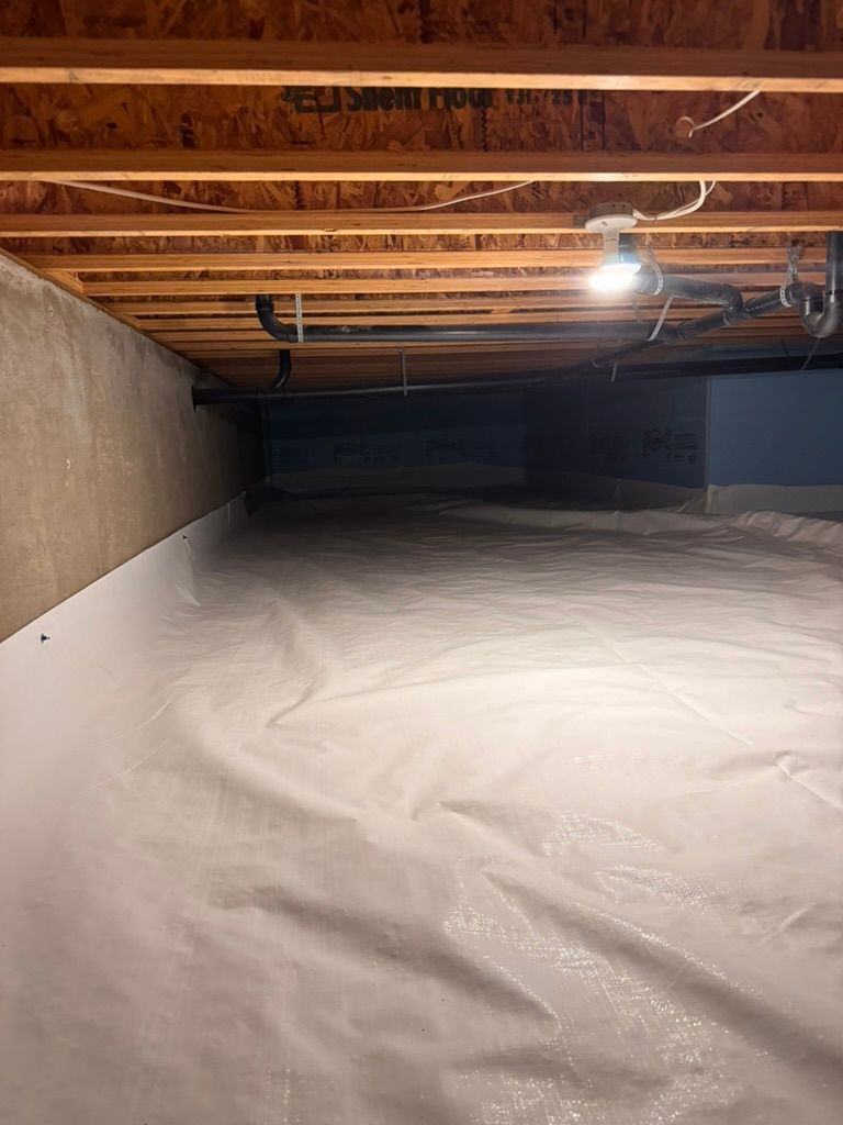 Crawlspace with exposed wooden beams, insulated left wall, bright light, and white vapor barrier floor covering.