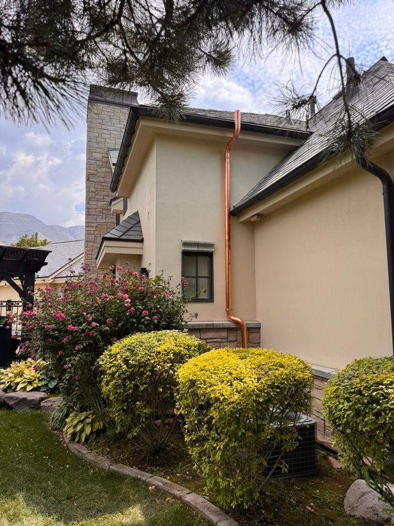 Exterior of a beige house with a red downspout, stone trim, and flowering shrubs in a garden