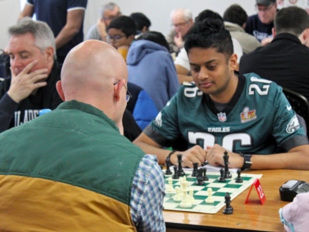 Two men playing chess at a crowded table, one in a green sports jersey, with other players in the background