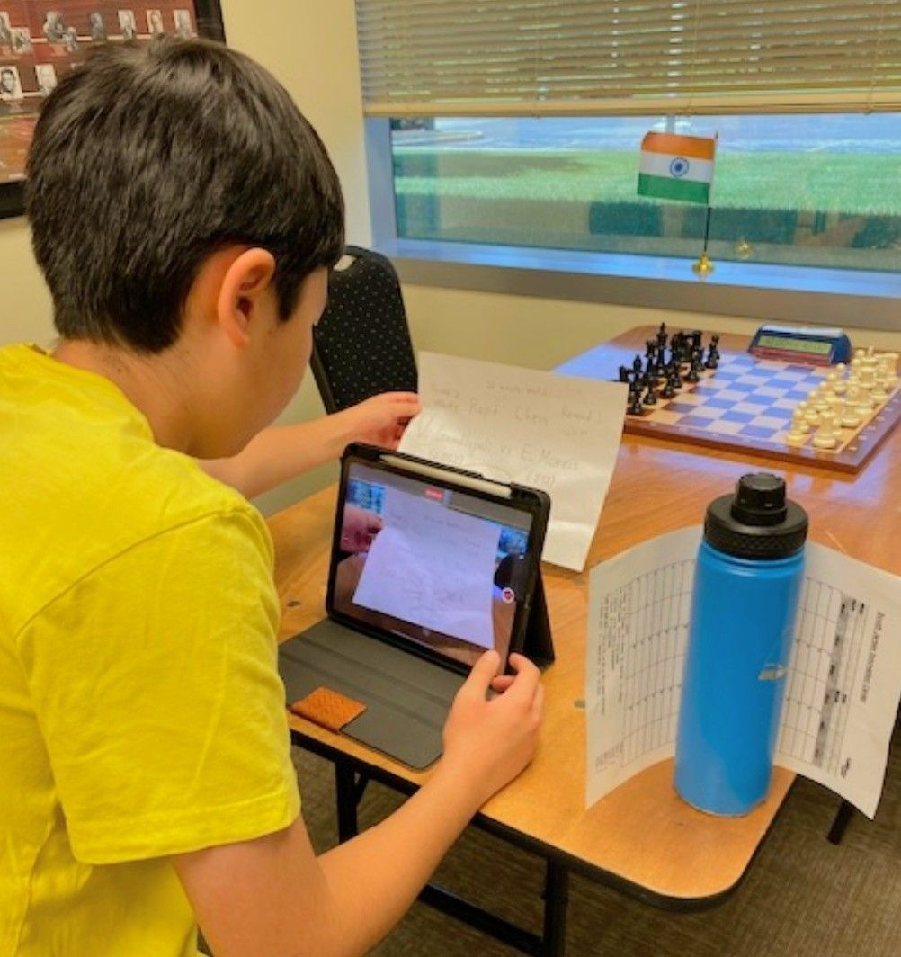 Student using a tablet at a desk to photograph a chessboard with the Indian flag on a classroom window.