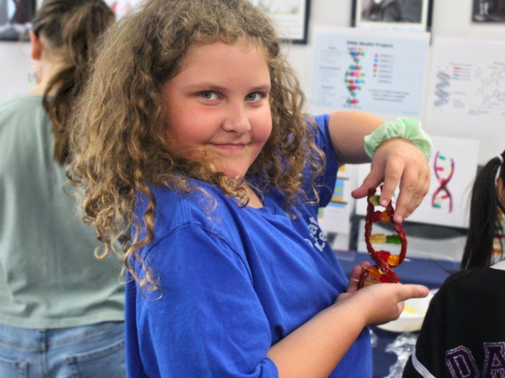 Child in a blue shirt smiling while holding colorful bracelets at a craft table in a classroom.