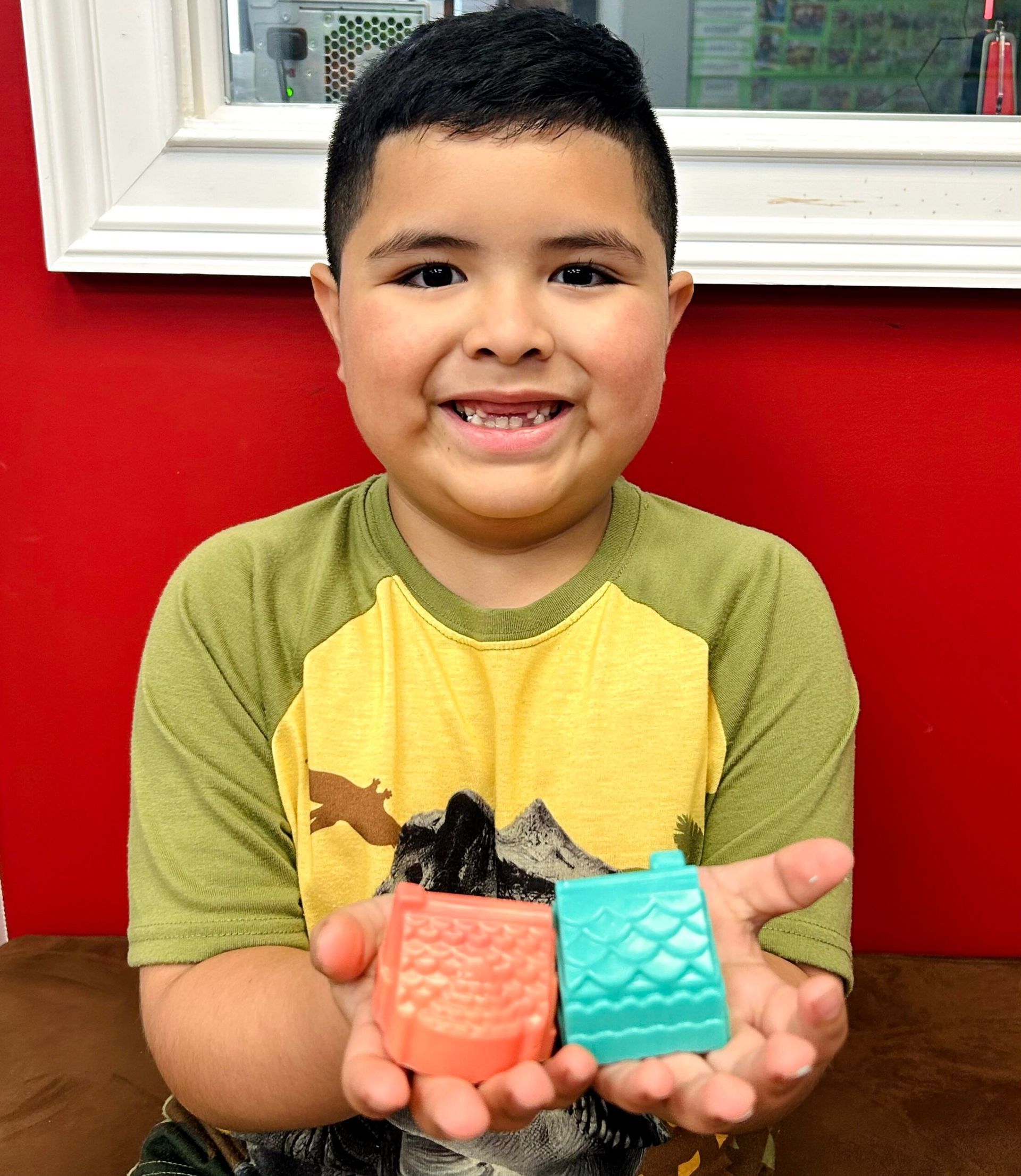 Child smiling and holding two colorful silicone items against a red wall