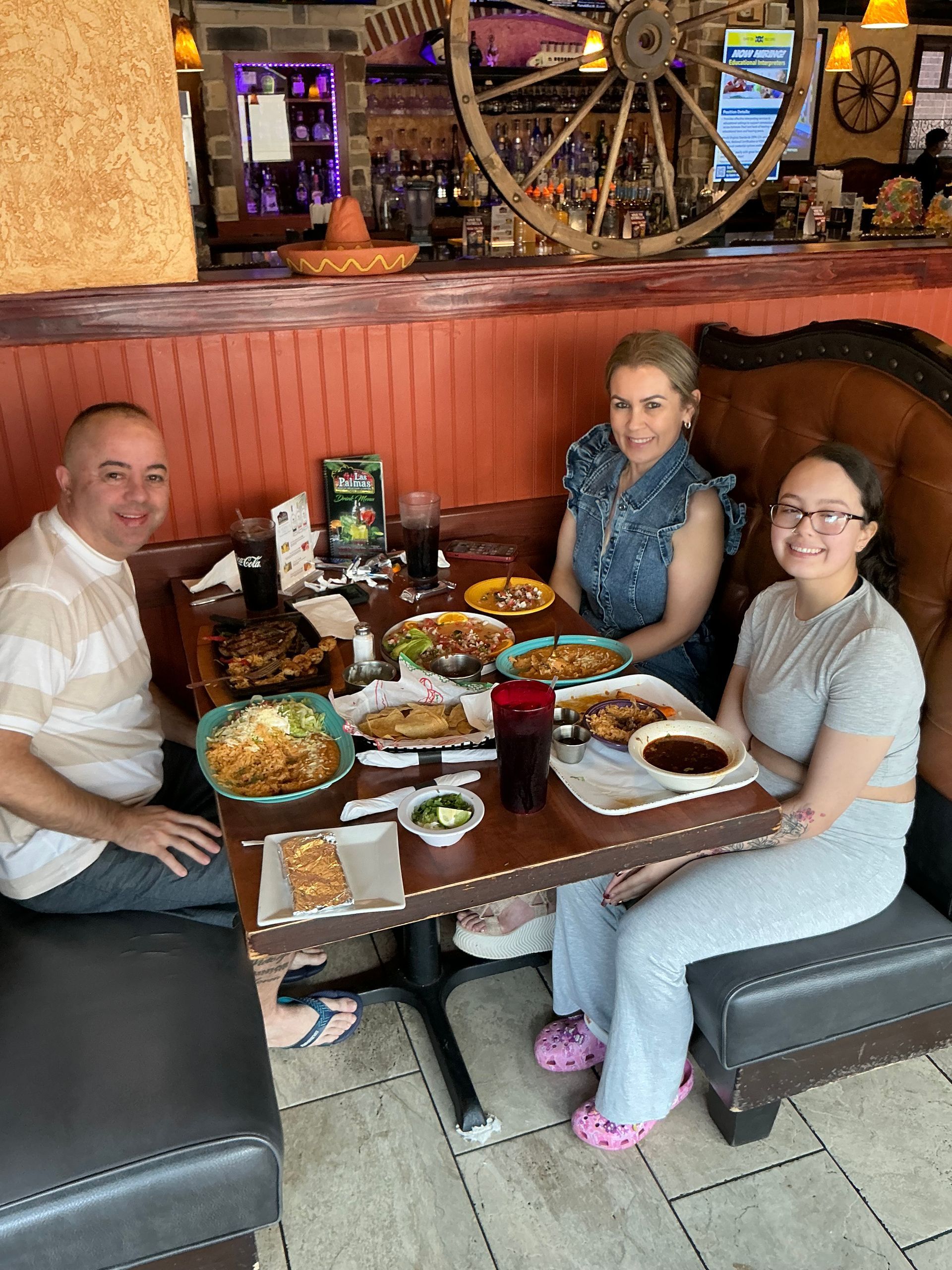 Three people seated at a restaurant table with food, including tacos and fried items. Brick wall backdrop.