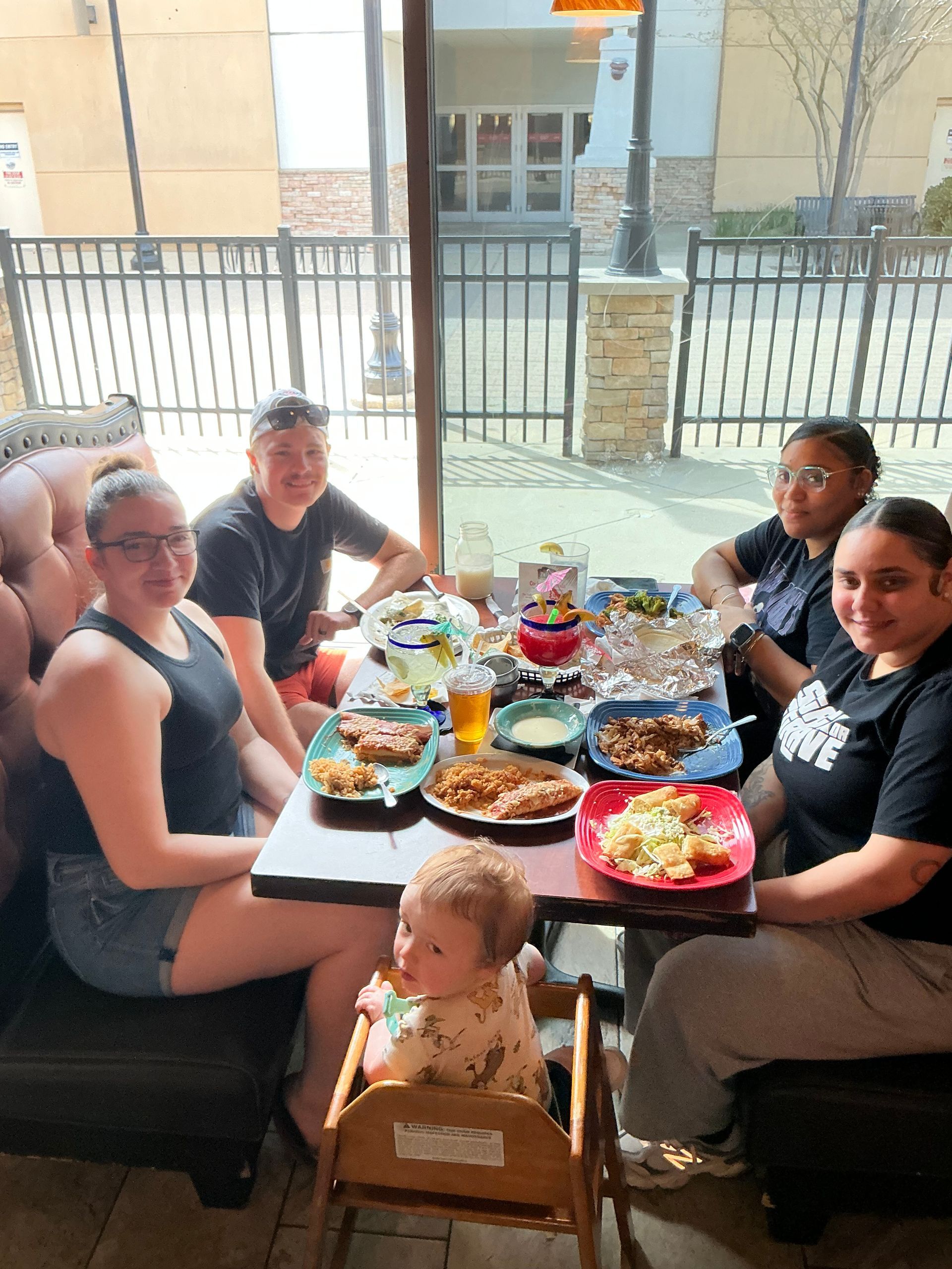 Group of people at a restaurant table with food, including a baby in a high chair. Natural light.