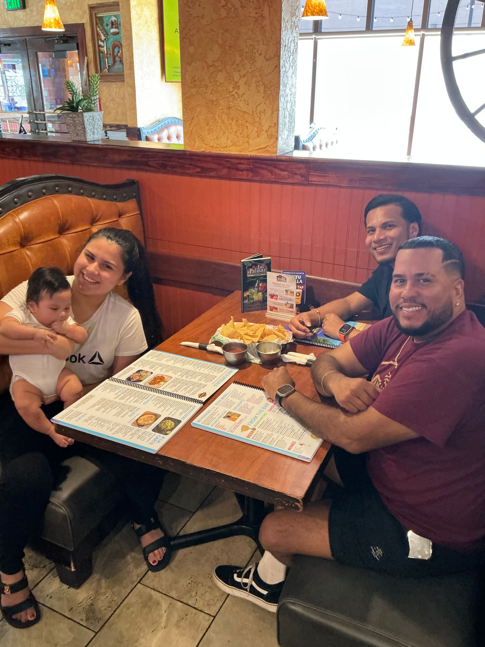 Four people sit at a restaurant table with a baby. They are smiling, and food and menus are present.