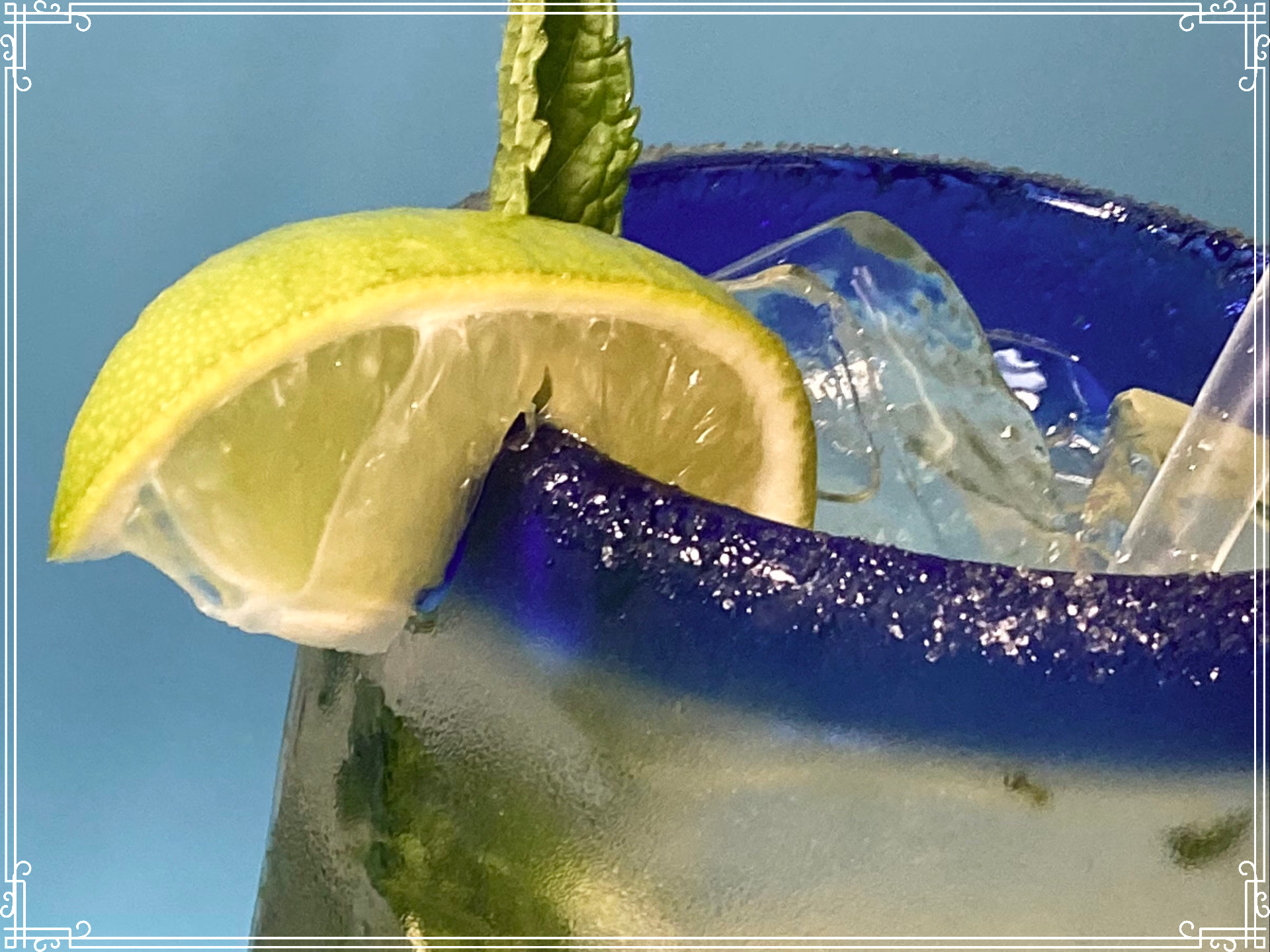 Close-up of a margarita glass with blue rim, lime slice, ice, and mint sprig against a blue background.