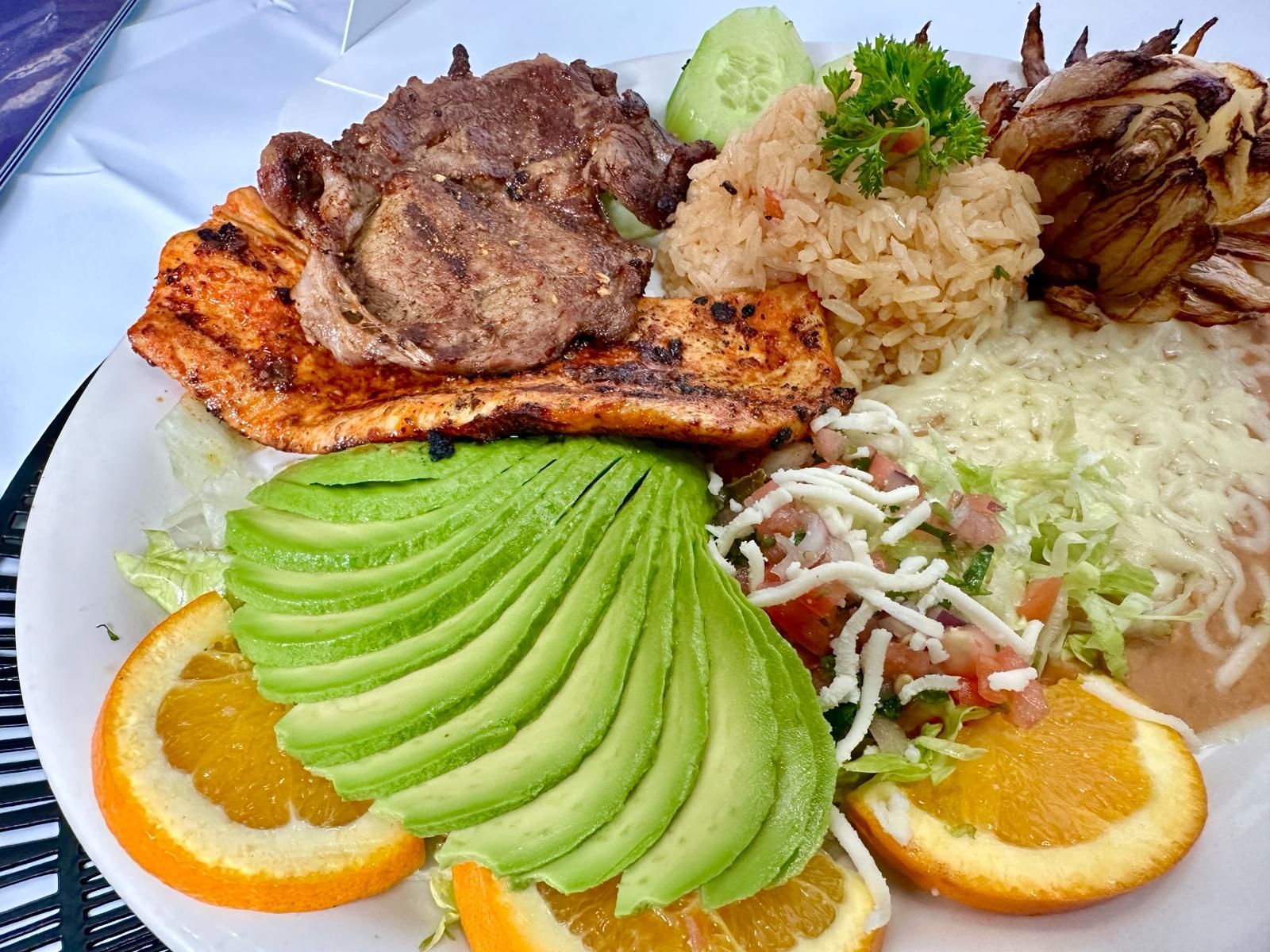 Plate of grilled chicken and steak, rice, beans, avocado slices, orange slices, and onions.