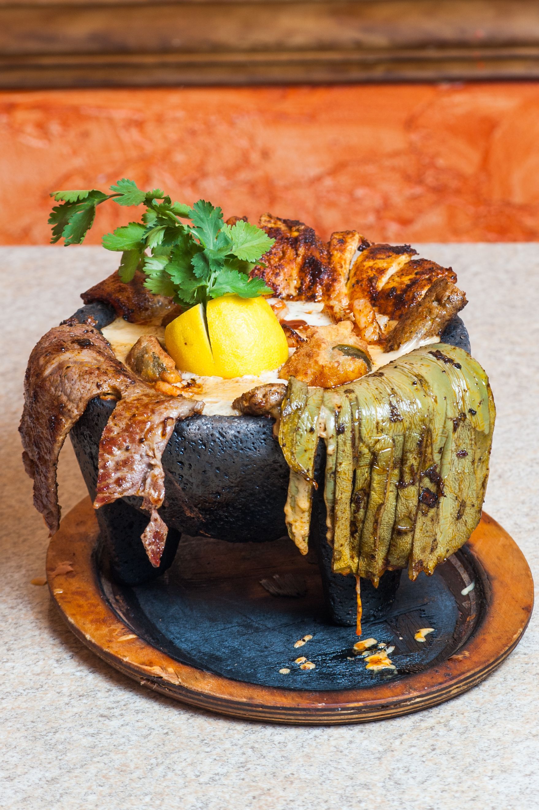 Molcajete dish: grilled meat, cheese, peppers, and lemon in a stone bowl, on a wooden plate.