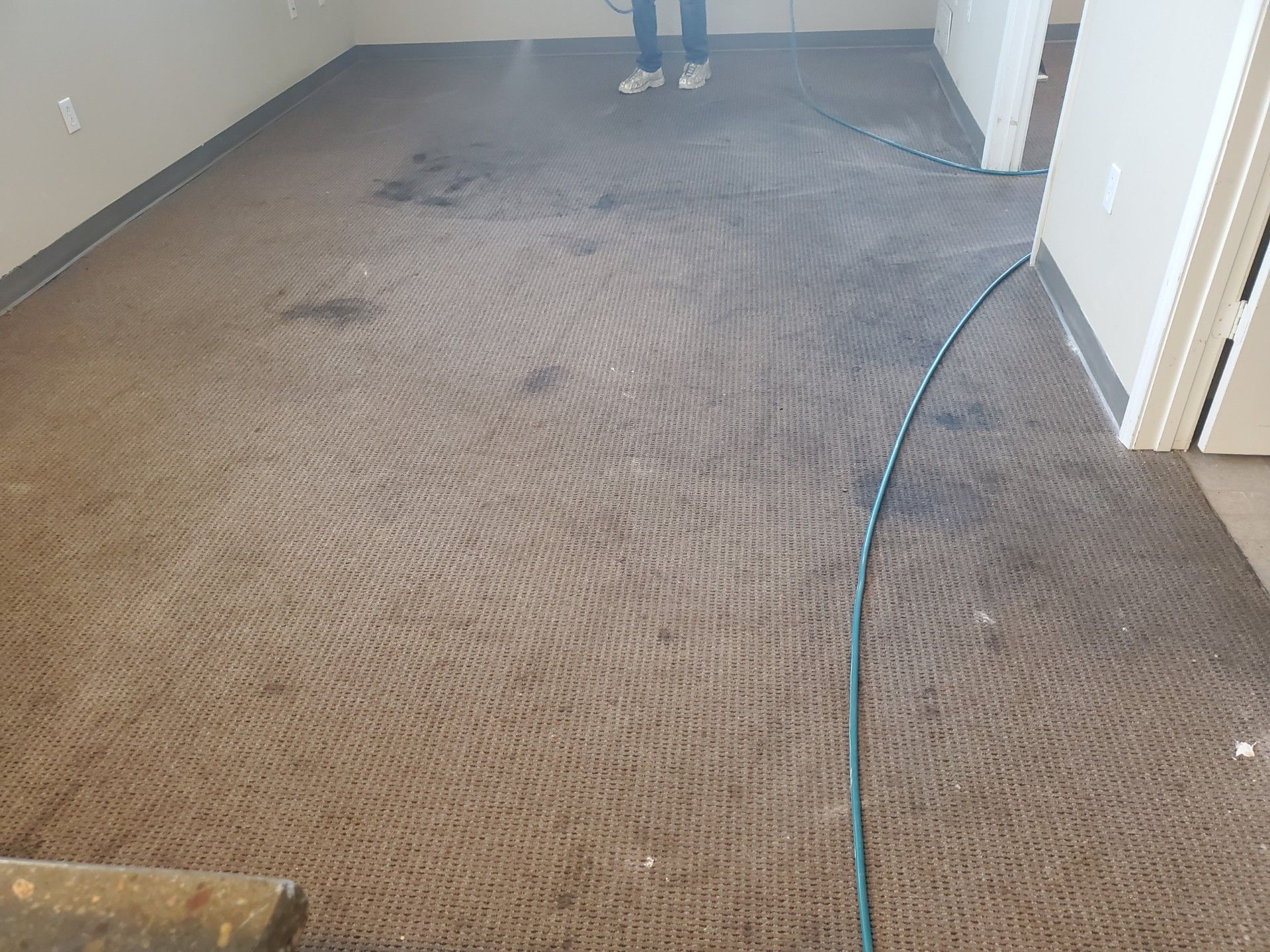 Carpeted room with visible dirt and footprints, a hose along the right wall, and a person standing at the far end