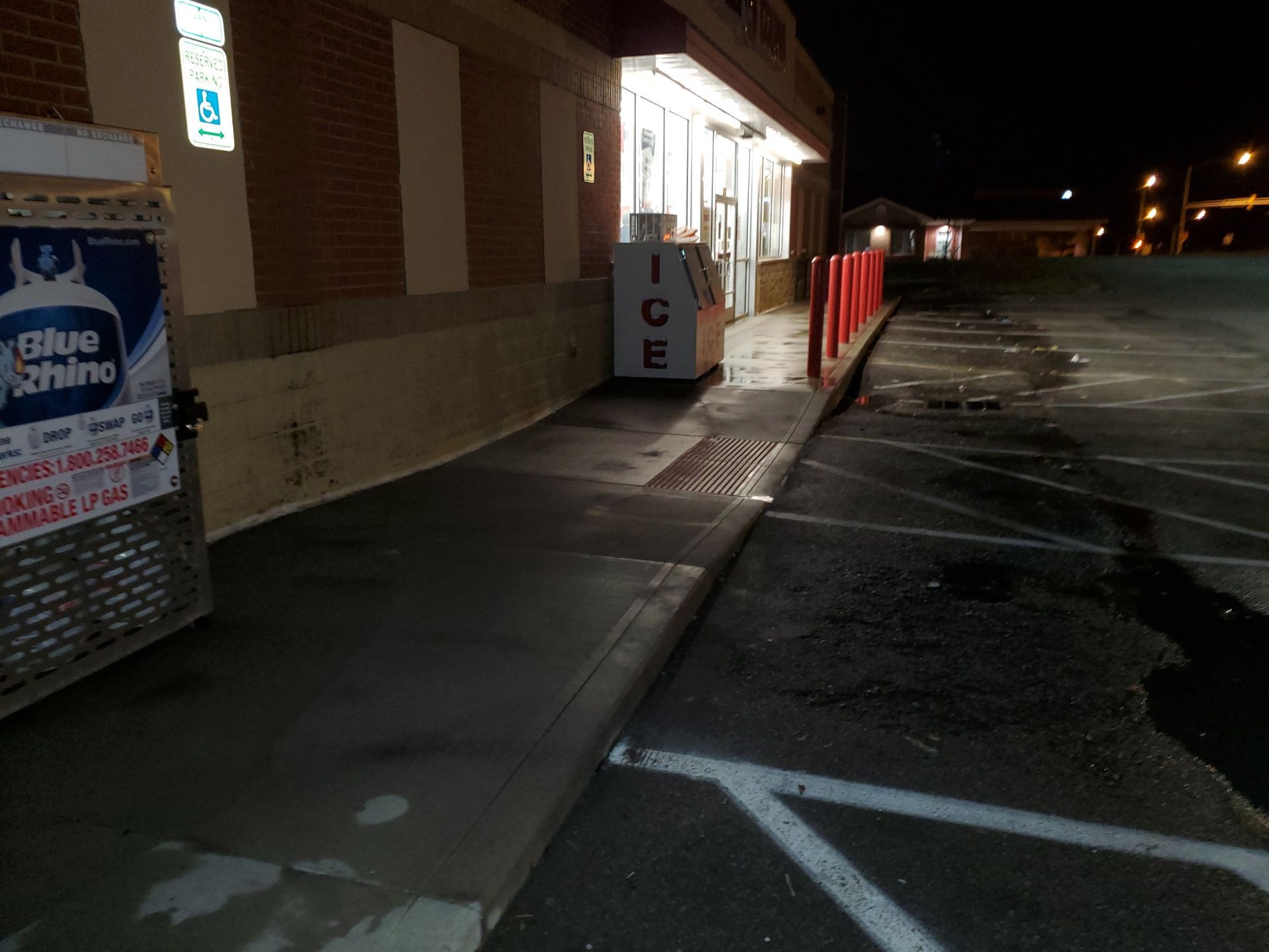 Nighttime parking lot beside a store, with a sidewalk, ice machine, and empty spaces.