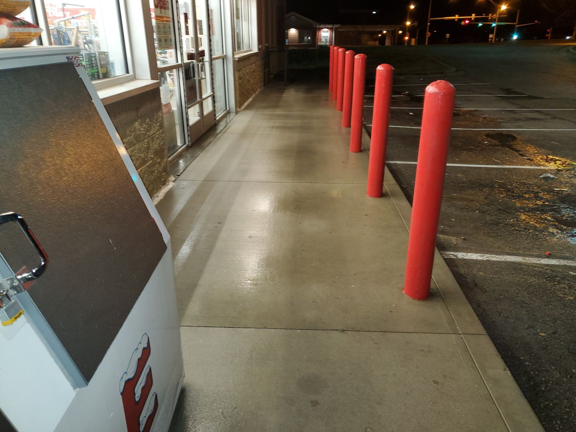 Storefront sidewalk at night with red bollards beside an empty parking lot
