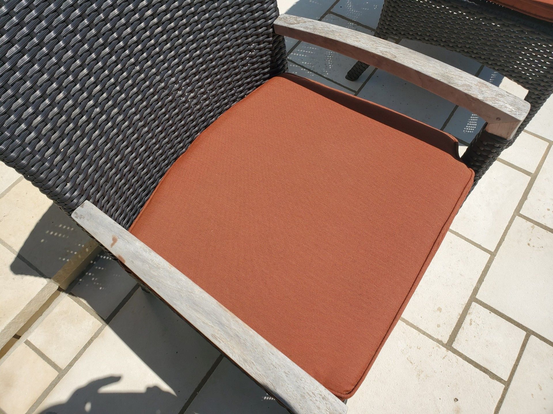 Outdoor chair with orange cushion, leaning against a woven black wall on a tiled patio.