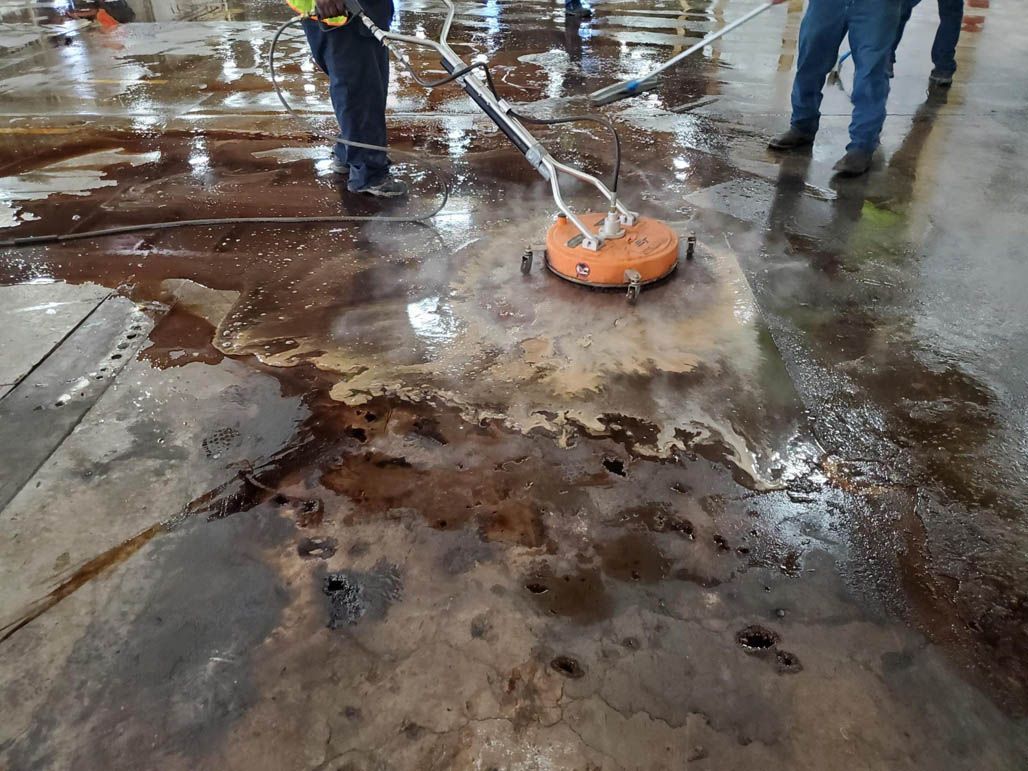 Workers pressure-washing a warehouse floor with an orange surface cleaning machine