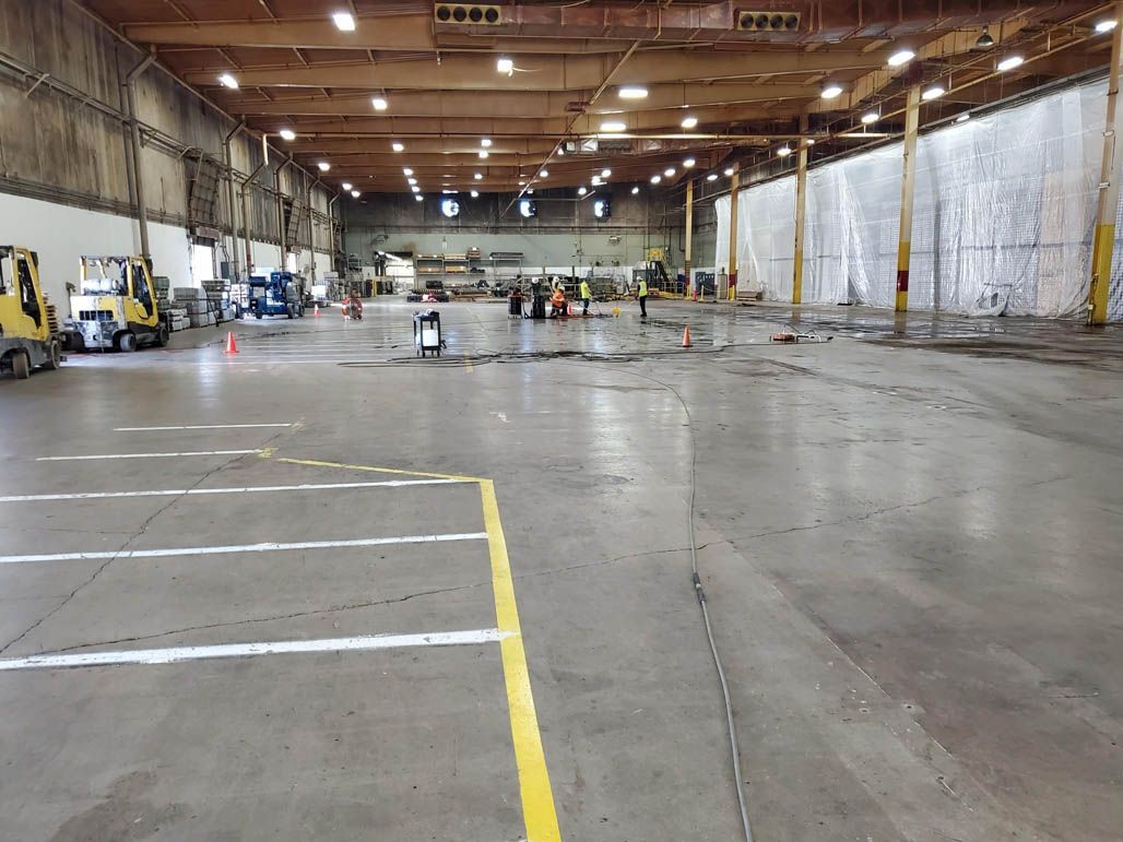 Large empty warehouse interior with a freshly power washed concrete floor, overhead lights, and parked equipment along the sides