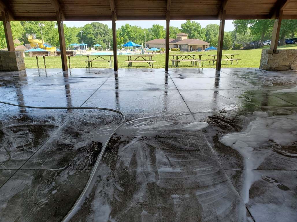 Covered concrete pavilion with wet floor and puddles overlooking a grassy park with picnic tables and blue umbrellas