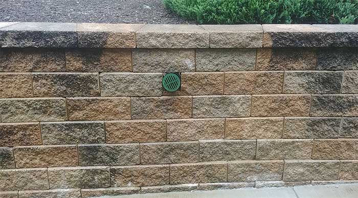 Stone retaining wall with a small green circular vent and greenery above it.