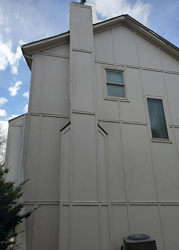 Exterior wall of a beige house with a chimney, windows, and a dark trash bin in the foreground.
