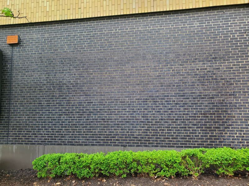 After graffiti was removed from a brick building wall with black siding above, orange light at left, and green shrubs along the bottom