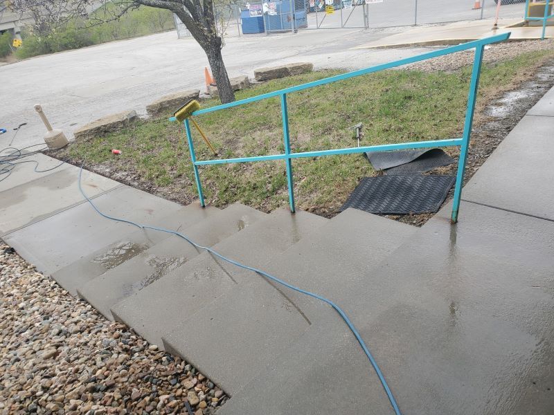 Damaged sidewalk ramp with blue handrails and cracked concrete beside a road