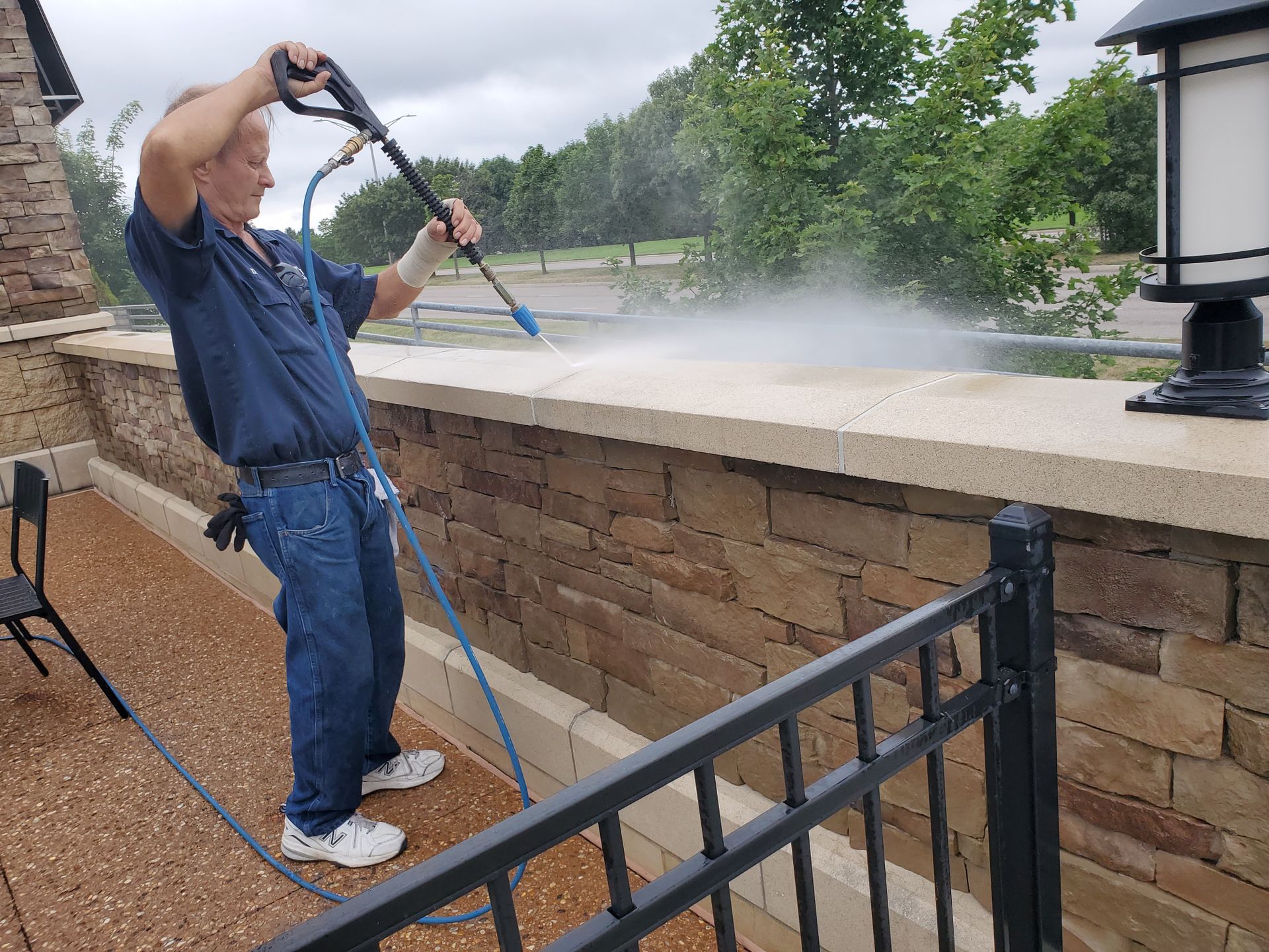 Worker power-washing a stone wall patio beside a black railing, with mist spraying across the outdoor area