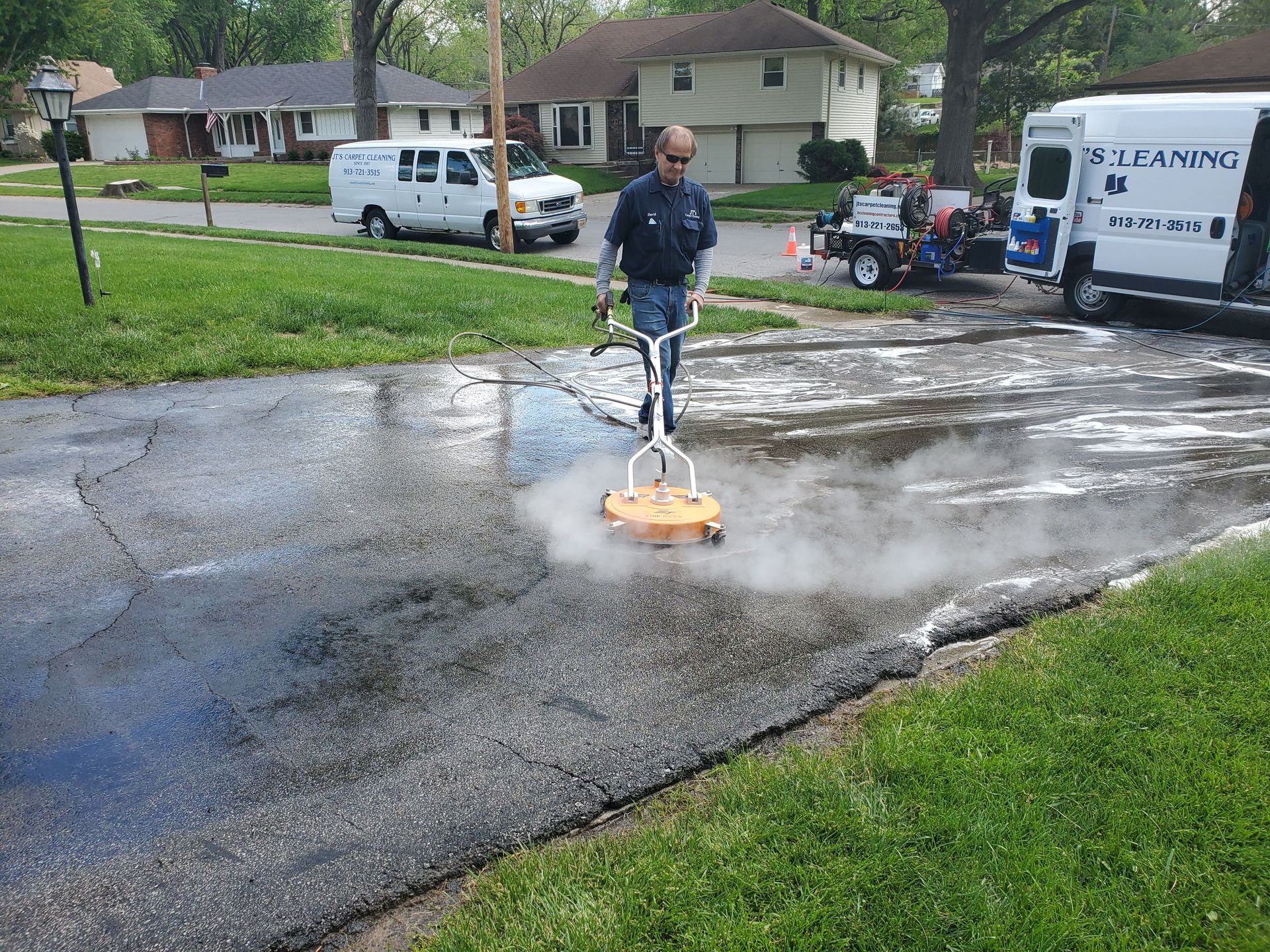 Worker pressure washing a driveway in a suburban neighborhood, with a white truck nearby.