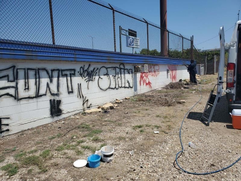 Prepping to clean a graffiti-covered wall beside a dirt lot, with scattered buckets.