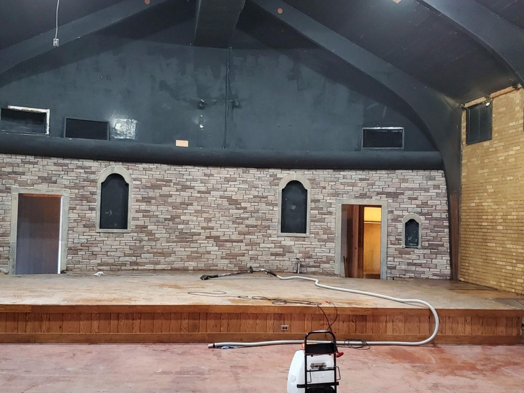 Empty indoor stage with stone backdrop, arched windows, wooden floor, and brick walls under a dark ceiling