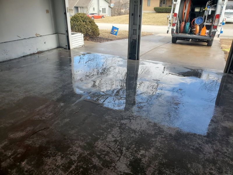 Wet concrete floor in a garage with standing water and a view of the driveway outside.