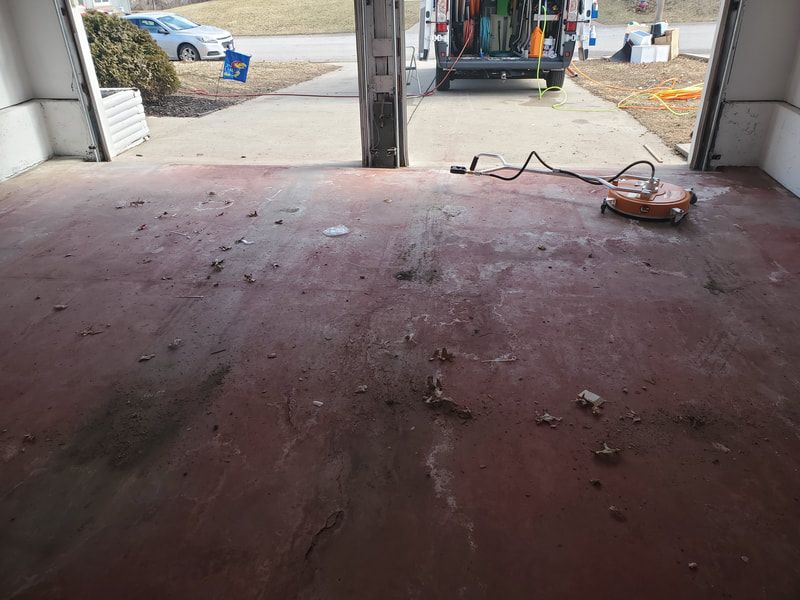 Dusty red garage floor with debris, a lawn tool, and an open garage door to a driveway.