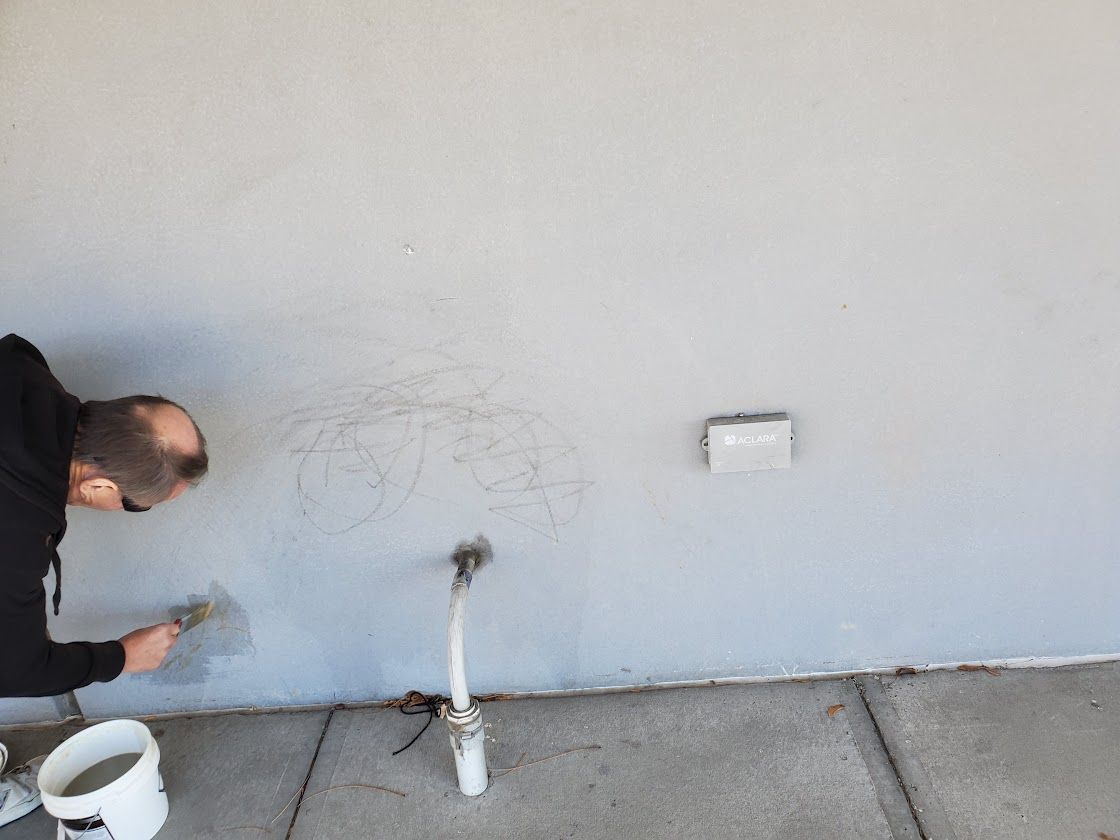 Worker scraping graffiti from a white wall near a pipe and bucket outside.