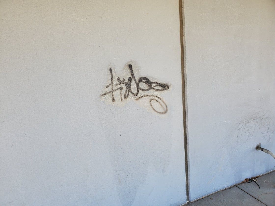 White wall with a small black graffiti tag near a vertical seam and a cable at the lower right.