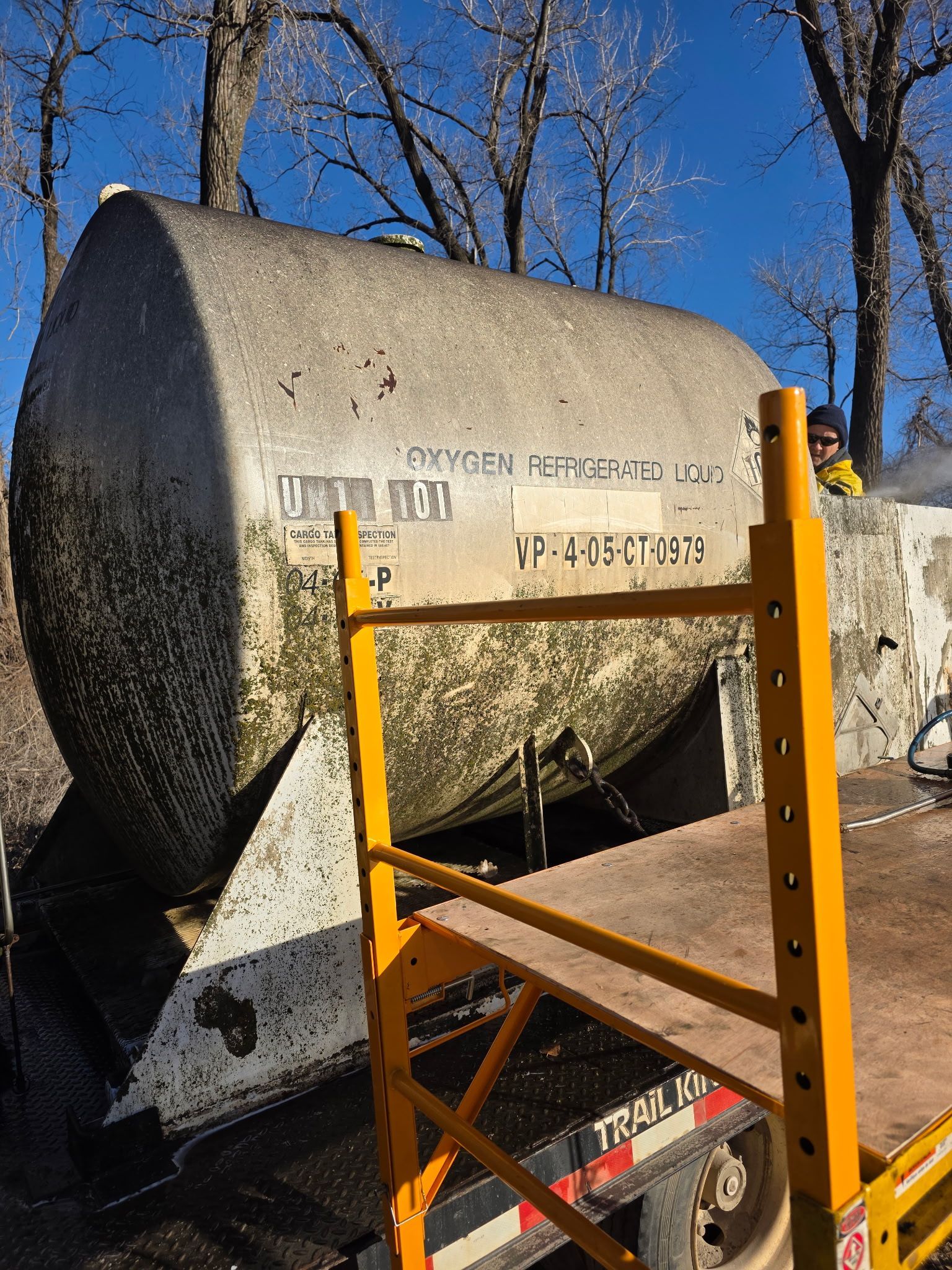 Before cleaning a large metal tank, with a yellow ladder in front, outdoors under a blue sky.