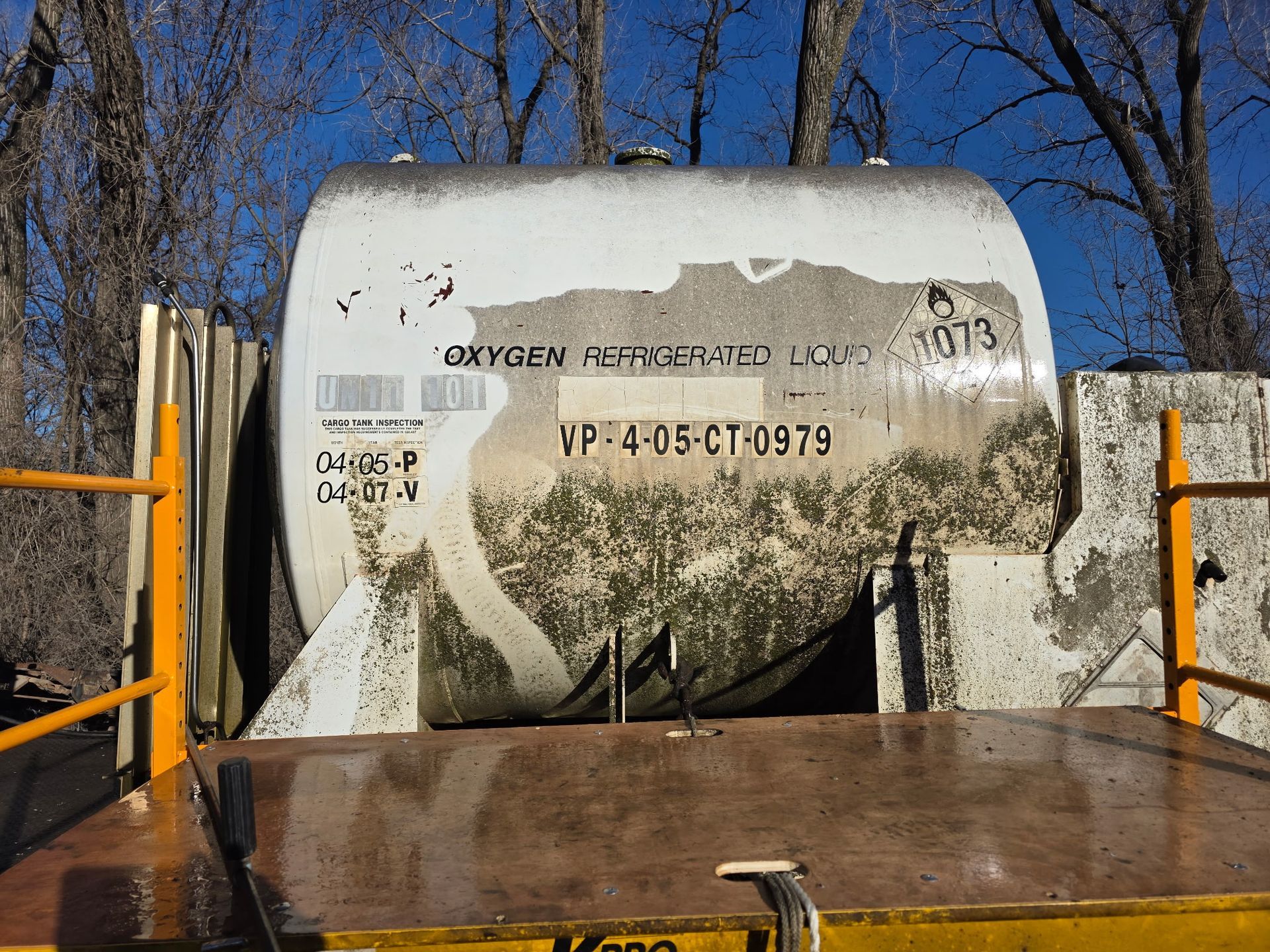 Before cleaning a white industrial tank with faded markings on a flatbed trailer outdoors among bare trees under a blue sky