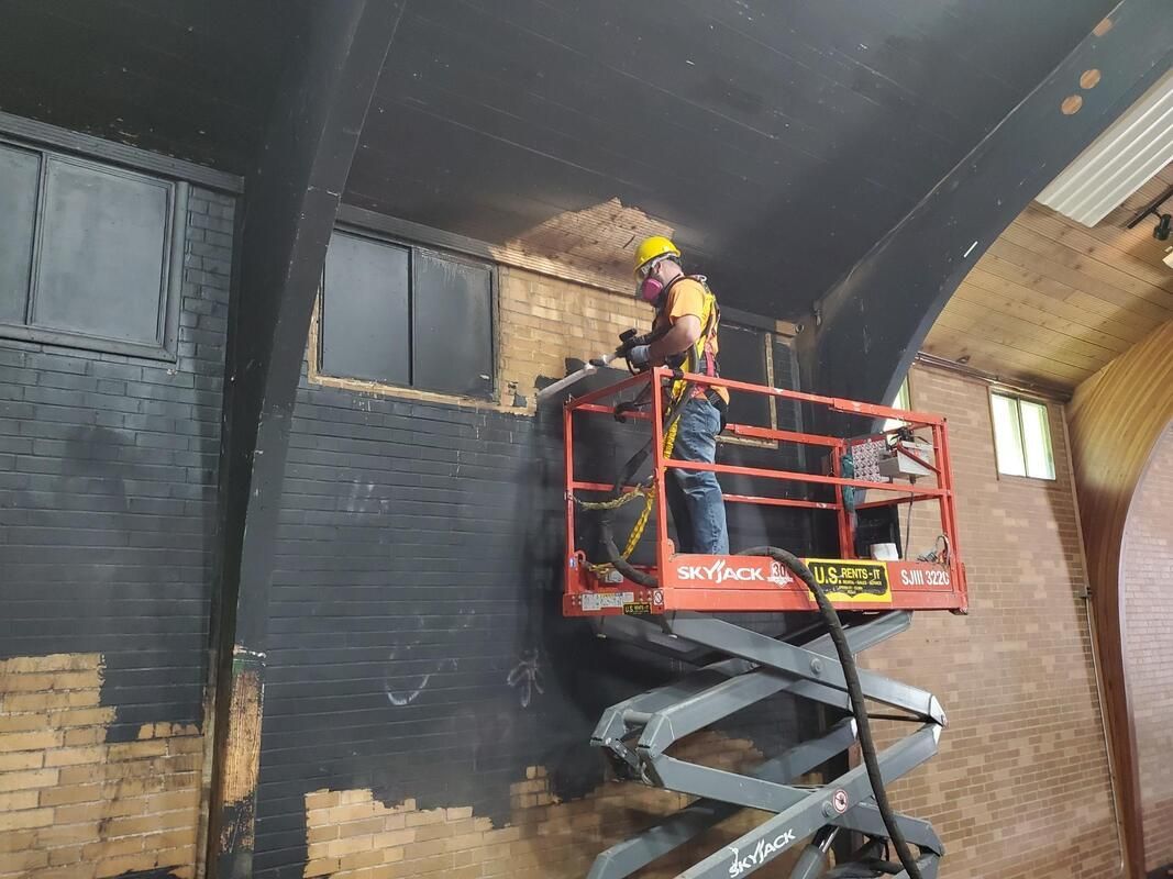 Worker in yellow hard hat on a red scissor lift painting a dark interior wall.