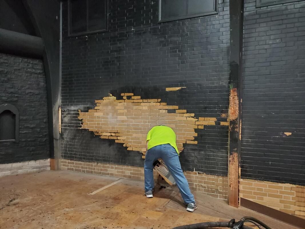 Worker in a neon green shirt kneels near a black brick wall, patching a large area of exposed brick.
