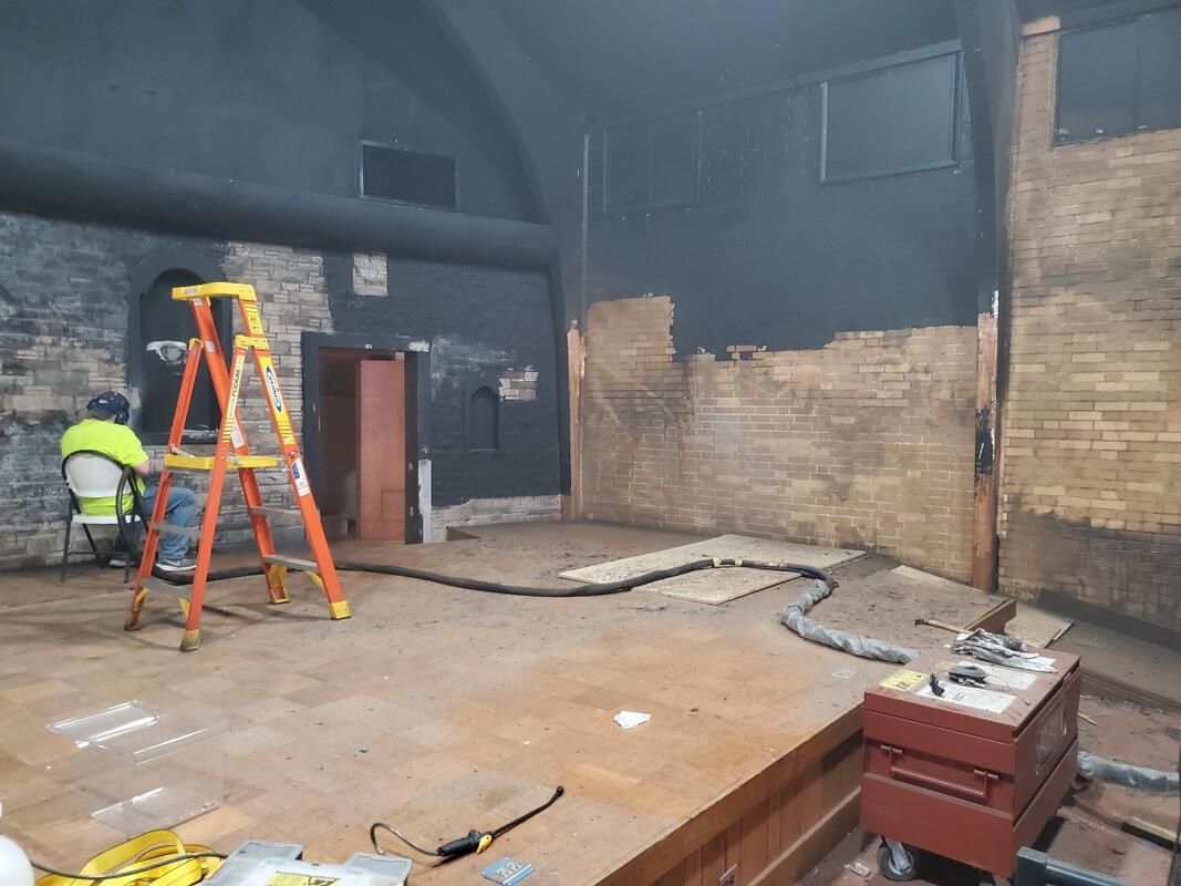 Fire-damaged room with two workers near a ladder, charred walls, and debris on the floor.