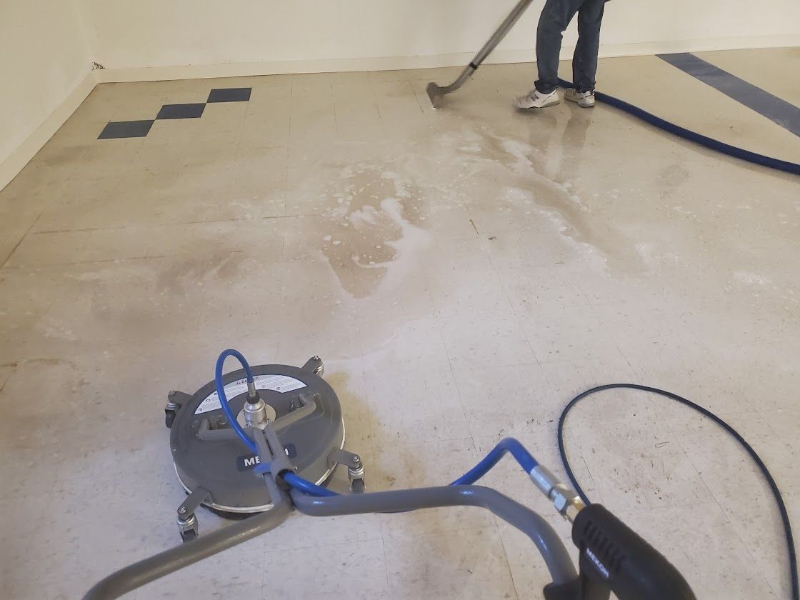 Person cleaning a beige floor with a floor buffer and hose in a room with dark tiles in the corner