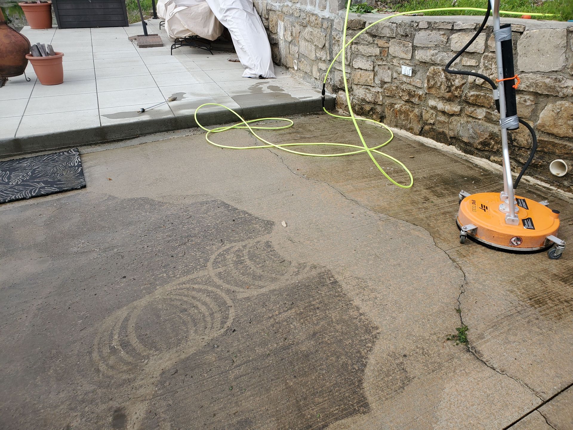 Orange floor scrubber cleaning a wet stone patio beside a stone wall and hose.