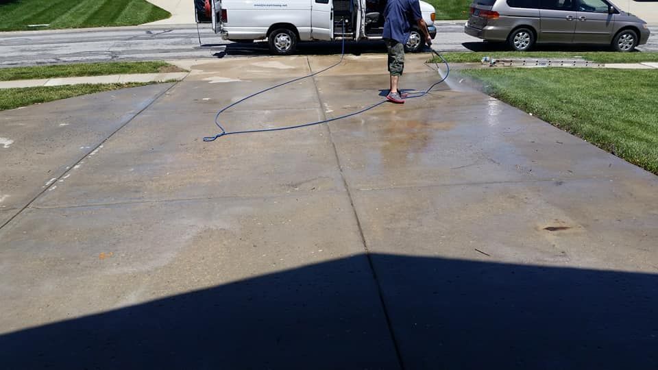 Person power-washing a wet driveway in front of parked cars and a green lawn