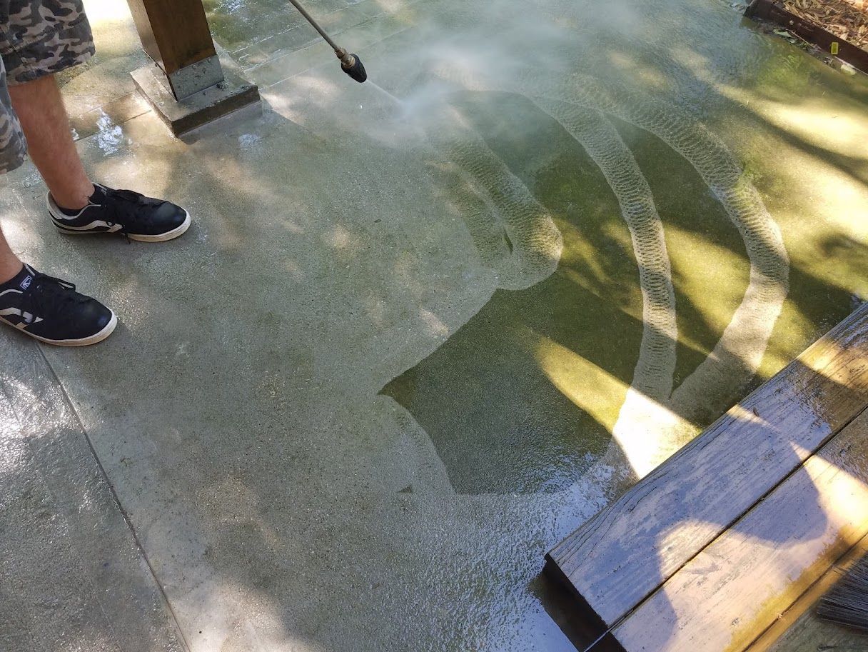 Two people standing on a wet paved surface with a broom and wooden bench nearby, in dappled sunlight