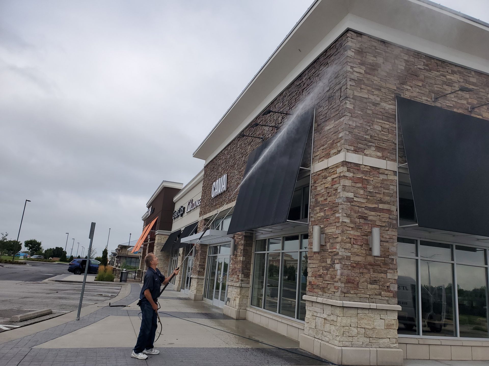 Person power washing outside a brick storefront with black awnings on a cloudy day