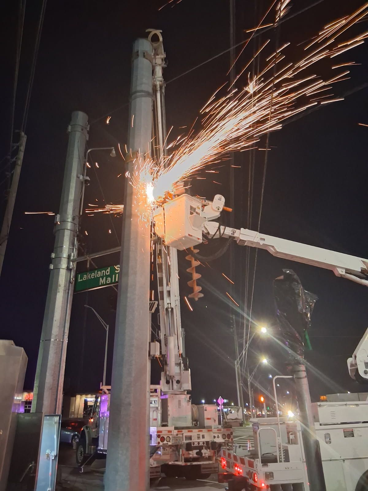 Worker cutting metal on a tall utility pole at night, with bright sparks flying and a boom truck nearby
