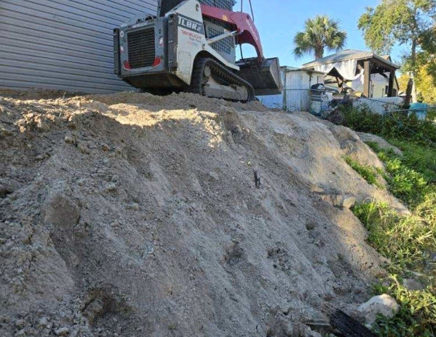 A white and red skid-steer loader sits at the top of a dirt embankment next to a building.