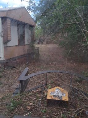 Abandoned outdoor scene with a small building and a rusted metal gate blocking a dirt path.