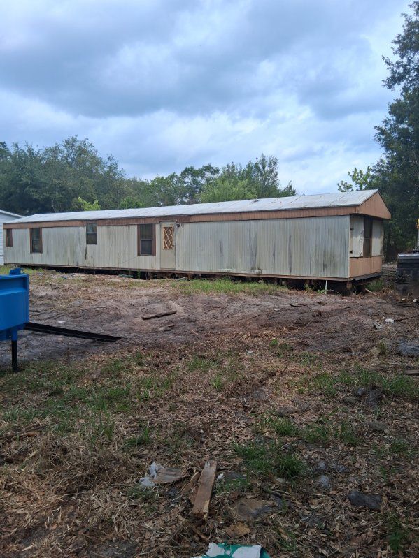 Long beige mobile home in a muddy lot under a cloudy sky, with trees and a blue dumpster nearby