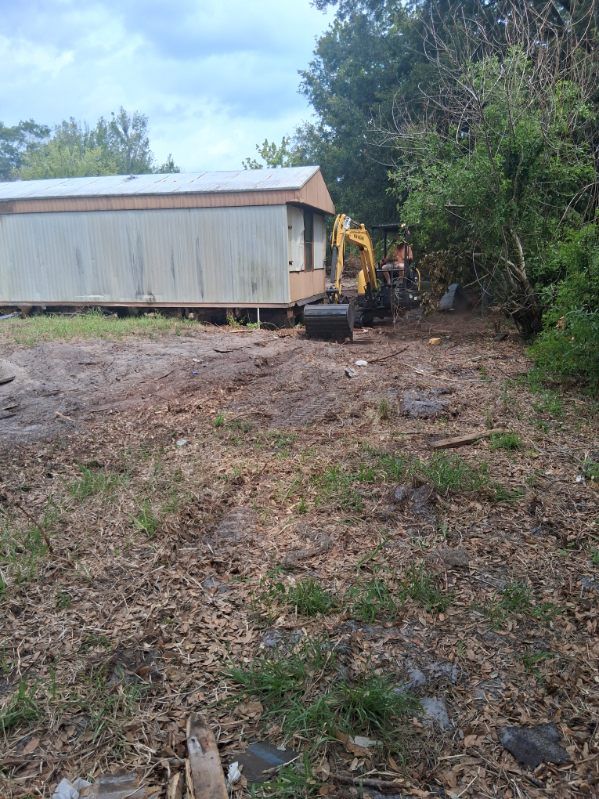 Wooded yard with a white shed and yellow excavator near trees and dirt ground