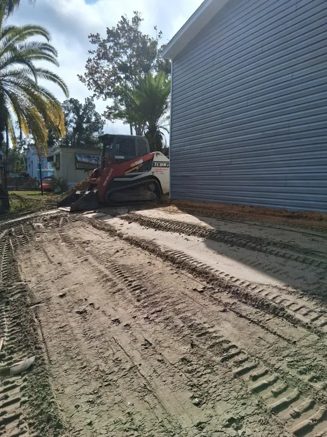 A red and white skid-steer loader sits next to the side of a grey-sided house on a dirt lot with tire tracks.