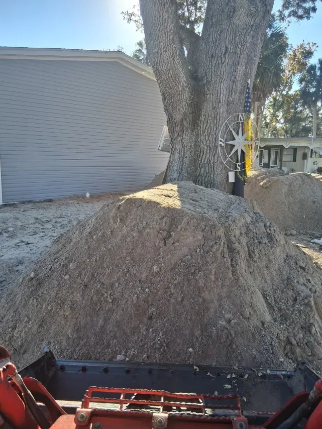 A view from the driver's seat of construction equipment looking at a large pile of dirt near a tree and a house.