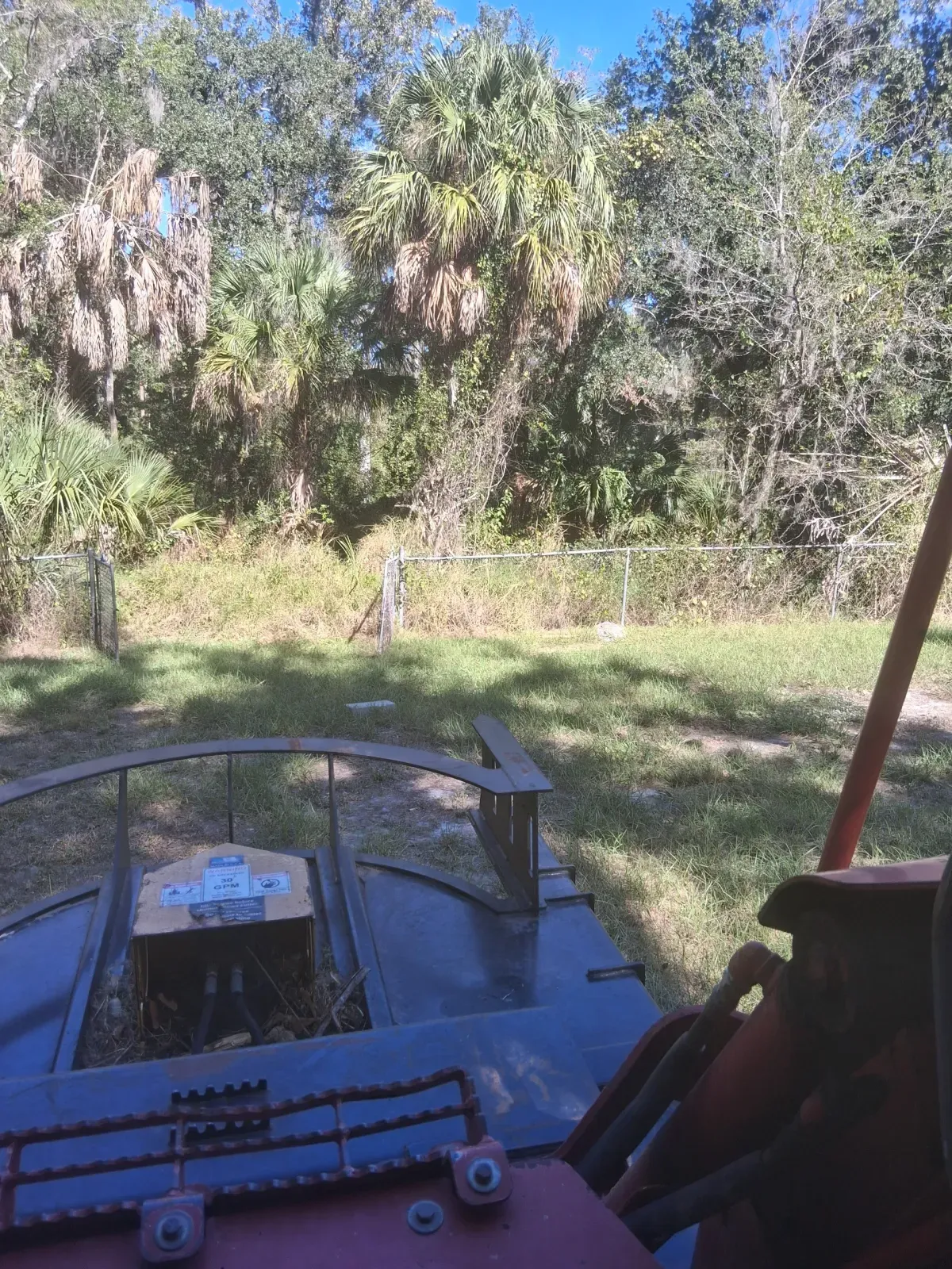 View from the driver's seat of a tractor looking out over a grassy field toward trees and a chain-link fence.